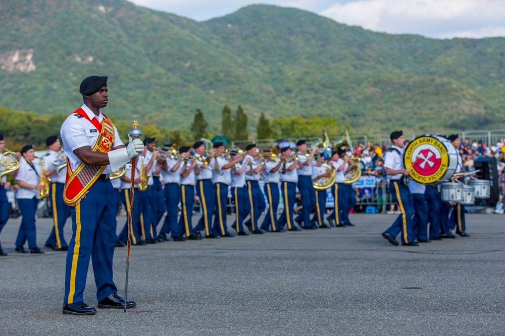 A U.S. Army military marching band, identified as the 'Eighth Army Band - Pacific Victors,' is performing an outdoor ceremony or parade. The band members are dressed in formal white dress uniforms with blue trousers and gold trim, led by a drum major in the foreground holding a ceremonial mace. The event appears to take place at a military installation or public venue with green mountainous terrain visible in the background.