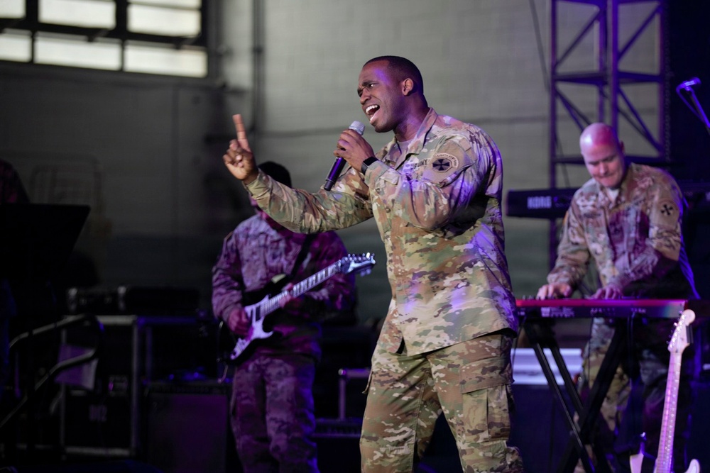 Military personnel in camouflage uniforms are performing a live musical concert on a stage. The lead performer is singing into a microphone while gesturing with one finger raised, accompanied by bandmates playing electric guitar and keyboard. The setting appears to be an indoor venue or hangar with stage lighting.