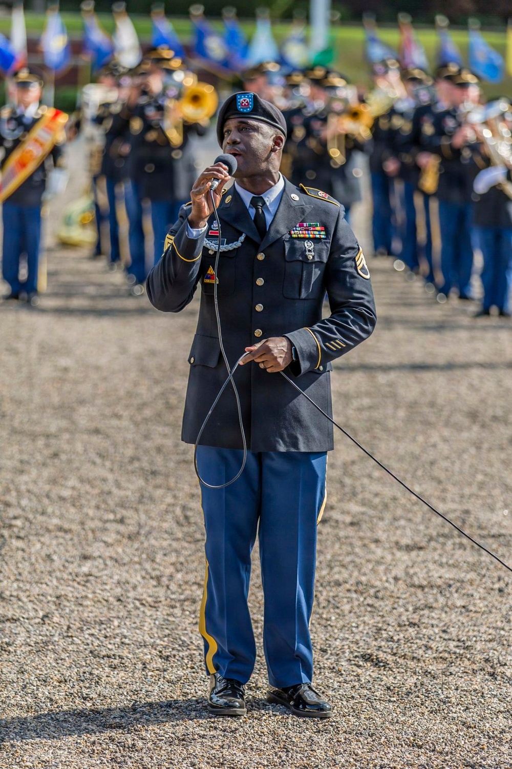 A U.S. Army soldier in dress uniform is performing as a vocalist at a formal military ceremony, singing into a handheld microphone while standing on a gravel parade ground. Behind him, a military band in dress blues is assembled in formation with brass instruments, and multiple unit guidon flags are visible in the background. The setting appears to be an outdoor military parade or ceremonial event.