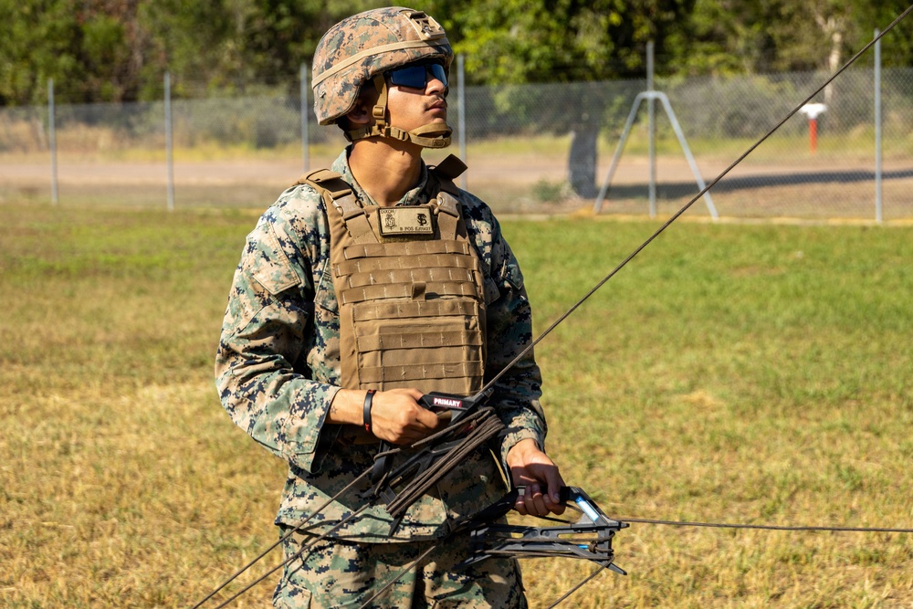 A U.S. Marine in full combat gear, including MARPAT digital camouflage uniform and plate carrier vest, holds what appears to be a compound bow or crossbow-related equipment with cables/strings visible. The setting is an outdoor grassy field with a chain-link fence and trees in the background, suggesting a training range environment. The label 'PRIMARY' is visible on the equipment being held.