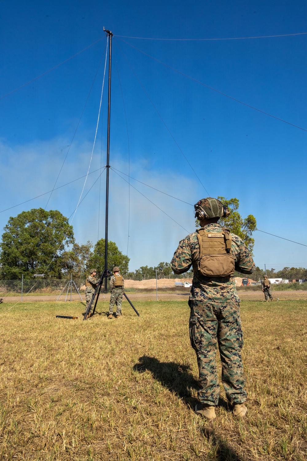 U.S. Marines in MARPAT camouflage uniforms are erecting a tall communications antenna mast in an open field. The foreground Marine, identified by name tape as 'SANDOVAL,' wears a tactical vest and hearing protection while observing the setup. The scene appears to be a field communications exercise at a military installation.