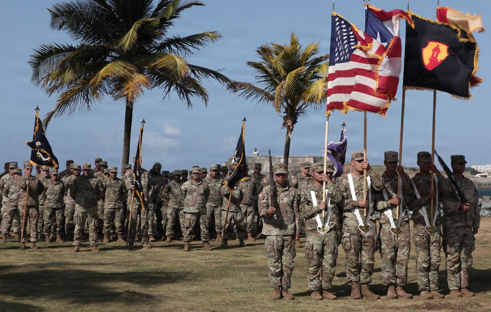 A formal military ceremony featuring U.S. Army soldiers in MultiCam/OCP camouflage uniforms standing in formation. A color guard in the foreground holds multiple flags including the U.S. flag, Puerto Rico flag, and a unit guidon with a yellow soldier silhouette emblem. The setting appears to be an outdoor parade ground near a historic fortification, with palm trees and coastal elements visible in the background.