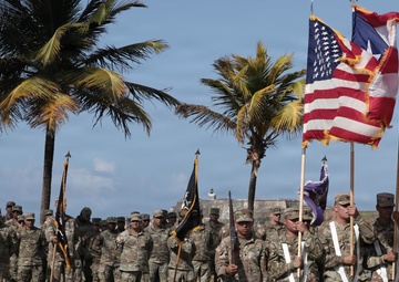 A formal military ceremony featuring U.S. Army soldiers in MultiCam/OCP camouflage uniforms standing in formation. A color guard in the foreground holds multiple flags including the U.S. flag, Puerto Rico flag, and a unit guidon with a yellow soldier silhouette emblem. The setting appears to be an outdoor parade ground near a historic fortification, with palm trees and coastal elements visible in the background.
