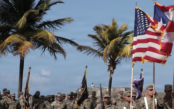 A formal military ceremony featuring U.S. Army soldiers in MultiCam/OCP camouflage uniforms standing in formation. A color guard in the foreground holds multiple flags including the U.S. flag, Puerto Rico flag, and a unit guidon with a yellow soldier silhouette emblem. The setting appears to be an outdoor parade ground near a historic fortification, with palm trees and coastal elements visible in the background.