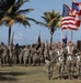 A formal military ceremony featuring U.S. Army soldiers in MultiCam/OCP camouflage uniforms standing in formation. A color guard in the foreground holds multiple flags including the U.S. flag, Puerto Rico flag, and a unit guidon with a yellow soldier silhouette emblem. The setting appears to be an outdoor parade ground near a historic fortification, with palm trees and coastal elements visible in the background.
