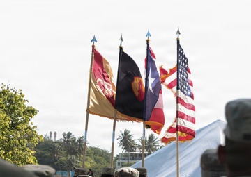 A military ceremony is taking place outdoors, featuring soldiers in U.S. Army camouflage uniforms (ACU/OCP pattern) standing in formation. Multiple flags are displayed including the United States flag, what appears to be the Puerto Rico flag, and U.S. Army unit guidons. The setting appears to be a tropical location based on the visible palm trees in the background.