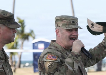 Two U.S. Army soldiers in OCP (Operational Camouflage Pattern) uniforms are participating in what appears to be a military ceremony or award presentation at an outdoor coastal location. One soldier is speaking into a microphone while holding up an engraved machete or large knife, likely a commemorative gift or award. The setting appears to be a beach or waterfront area with palm trees visible in the background.