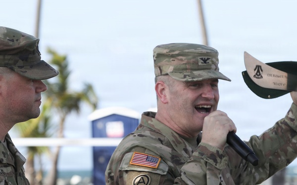 Two U.S. Army soldiers in OCP (Operational Camouflage Pattern) uniforms are participating in what appears to be a military ceremony or award presentation at an outdoor coastal location. One soldier is speaking into a microphone while holding up an engraved machete or large knife, likely a commemorative gift or award. The setting appears to be a beach or waterfront area with palm trees visible in the background.