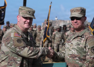 Two U.S. Army officers are shaking hands during a formal military ceremony while jointly holding a green official document folder bearing a government seal. They are standing in the foreground of a parade field with a large formation of soldiers in the background. The setting appears to be an outdoor military installation near a historic fortification structure.