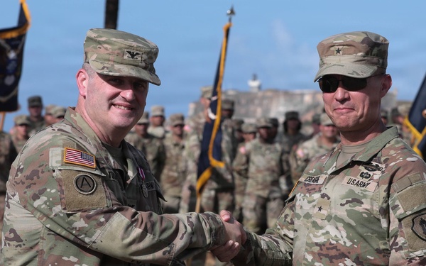 Two U.S. Army officers are shaking hands during a formal military ceremony while jointly holding a green official document folder bearing a government seal. They are standing in the foreground of a parade field with a large formation of soldiers in the background. The setting appears to be an outdoor military installation near a historic fortification structure.