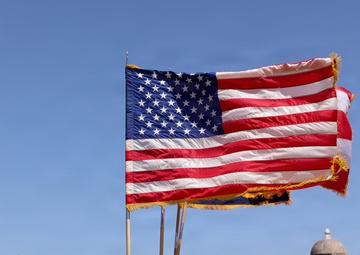 A U.S. Army color guard unit is conducting a formal military ceremony outdoors, holding large American flags with gold fringe on wooden staffs. The ceremony takes place on a grassy field adjacent to what appears to be a historic Spanish colonial fortification, likely Castillo San Felipe del Morro in San Juan, Puerto Rico. A senior soldier in camouflage uniform approaches the color guard formation from the right.