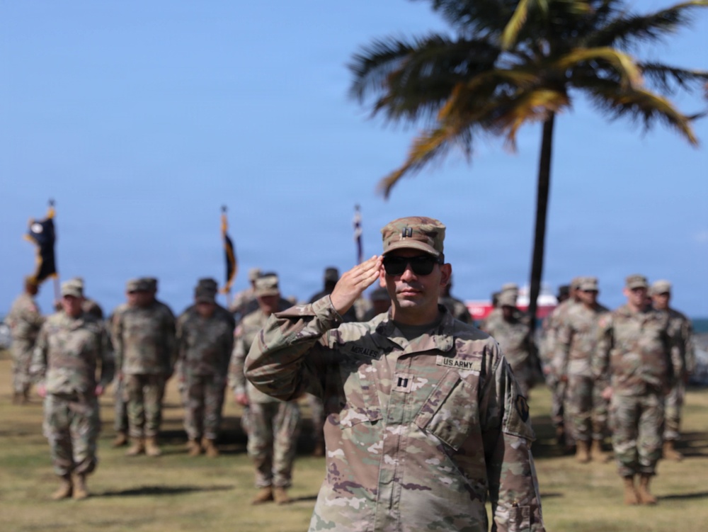 A US Army Captain is rendering a salute during what appears to be a formal military ceremony outdoors. The soldier in the foreground, identified by a name tape reading 'MORALES' and captain rank insignia, is in sharp focus while numerous other uniformed soldiers stand in formation behind him. The tropical setting, indicated by palm trees and blue sky, suggests a location such as Hawaii or Puerto Rico.