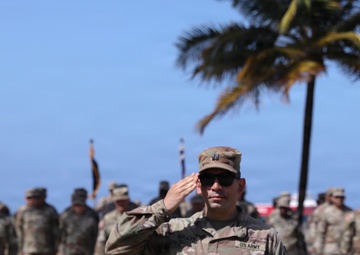 A US Army Captain is rendering a salute during what appears to be a formal military ceremony outdoors. The soldier in the foreground, identified by a name tape reading 'MORALES' and captain rank insignia, is in sharp focus while numerous other uniformed soldiers stand in formation behind him. The tropical setting, indicated by palm trees and blue sky, suggests a location such as Hawaii or Puerto Rico.