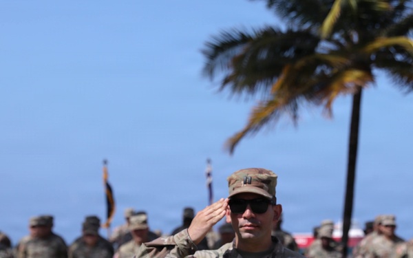 A US Army Captain is rendering a salute during what appears to be a formal military ceremony outdoors. The soldier in the foreground, identified by a name tape reading 'MORALES' and captain rank insignia, is in sharp focus while numerous other uniformed soldiers stand in formation behind him. The tropical setting, indicated by palm trees and blue sky, suggests a location such as Hawaii or Puerto Rico.
