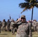A US Army Captain is rendering a salute during what appears to be a formal military ceremony outdoors. The soldier in the foreground, identified by a name tape reading 'MORALES' and captain rank insignia, is in sharp focus while numerous other uniformed soldiers stand in formation behind him. The tropical setting, indicated by palm trees and blue sky, suggests a location such as Hawaii or Puerto Rico.
