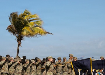 A military formation of soldiers in camouflage uniforms is performing a salute during a formal outdoor ceremony near a coastal waterfront. Multiple unit guidons and flags with gold fringe are being displayed, suggesting an official military event such as a change of command, activation, or awards ceremony. The setting appears to be a tropical coastal location with palm trees bending in the wind and a harbor visible in the background.