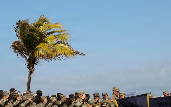 A military formation of soldiers in camouflage uniforms is performing a salute during a formal outdoor ceremony near a coastal waterfront. Multiple unit guidons and flags with gold fringe are being displayed, suggesting an official military event such as a change of command, activation, or awards ceremony. The setting appears to be a tropical coastal location with palm trees bending in the wind and a harbor visible in the background.