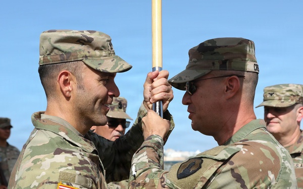 Two U.S. Army soldiers in OCP (Operational Camouflage Pattern) uniforms are engaged in a guidon transfer ceremony, likely a change of command. The soldier on the left wears an Airborne tab and Special Forces (crossed arrows) patch with a U.S. flag on his right sleeve. The ceremony appears to be taking place outdoors near a historic stone fortification, with a formation of soldiers visible in the background.