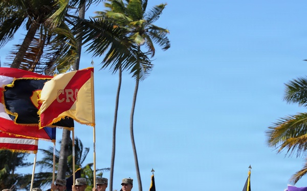 A formal military ceremony is taking place outdoors in a tropical setting, likely Hawaii, with soldiers in Army Combat Uniforms (ACUs) standing in formation. A color guard is prominently displayed at the front, carrying multiple flags including the United States flag and unit guidons. The backdrop features tall palm trees and a clear blue sky near what appears to be a coastline.