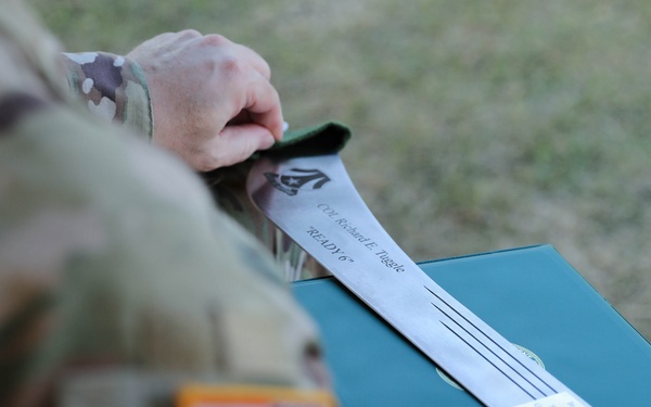 A U.S. Army soldier in OCP (Operational Camouflage Pattern) uniform is holding a green presentation/award case with a document or citation visible on top. The document is partially legible and references 'COL Richard E. Tuggle' and the nickname 'READY 6'. The soldier's sleeve displays a reverse U.S. flag patch and what appears to be a Third Army (ARCENT) shoulder sleeve insignia.