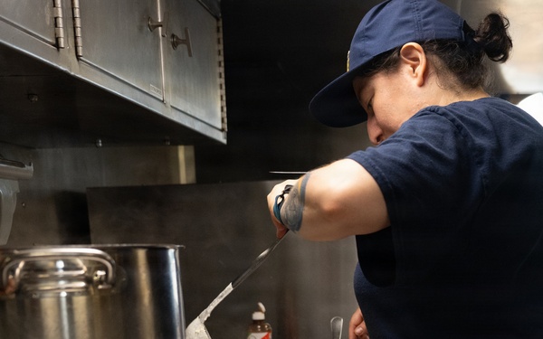 A person wearing a navy blue uniform and cap is working in a commercial or institutional kitchen, pouring liquid from a ladle into a large metal pan containing what appears to be a grain or rice dish. The setting appears to be a galley kitchen, possibly aboard a vessel or in a military/government facility, suggested by the compact stainless steel equipment and the uniform with 'STEADY' visible on the back. The image captures an active food preparation moment under overhead ventilation equipment.