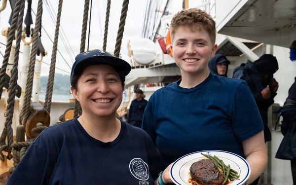 Two crew members in matching navy blue uniforms pose together on the deck of a tall ship, smiling at the camera. One crew member holds a white plate with a plated meal consisting of what appears to be a braised meat dish with green beans and sauce. The setting is a wet ship deck, suggesting rainy conditions, with rigging, ropes, and other crew members visible in the background.