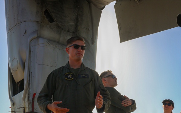 Two military aviators in olive green flight suits stand on a tarmac beneath a large military helicopter, appearing to brief or explain something to observers. A civilian woman wearing a lanyard/badge and a cap stands nearby, suggesting a public affairs or media event. The setting appears to be an airshow or base open house based on the visible aircraft and vehicles in the background.