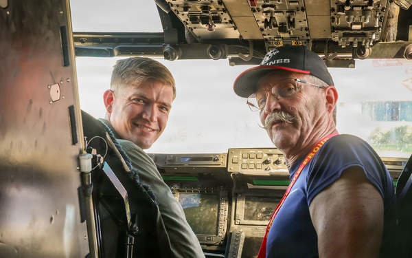 Two men are seated inside the cockpit of a military aircraft, posing for a photo. One man is wearing a green flight suit, suggesting he is a military aviator or crew member, while the other is a civilian visitor wearing a navy blue t-shirt, baseball cap, and a lanyard/badge. The cockpit features multiple avionics displays, control panels, and instrumentation consistent with a large military aircraft.