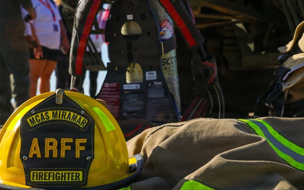 A yellow firefighter helmet and turnout gear are laid out on a tarmac surface at MCAS Miramar. The helmet is marked 'ARFF FIREFIGHTER' indicating Aircraft Rescue and Fire Fighting personnel. The gear belongs to a firefighter whose name appears to read 'ALEJANDRO' on the turnout pants.