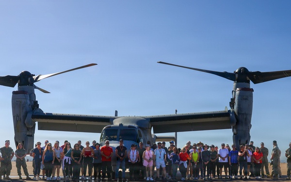 A large group of approximately 40-50 people, including military personnel in uniform and civilians in casual clothing, pose for a group photograph in front of a Bell Boeing V-22 Osprey tiltrotor aircraft on a military airfield tarmac. The aircraft's rotors are in the horizontal (airplane mode) position, with both large prop-rotors clearly visible extending from each wingtip. The setting appears to be a formal visit or public affairs event at a military installation.