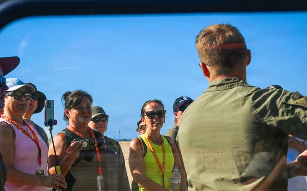 The image is captured through a vehicle window or mirror, showing a military personnel in olive green flight suit briefing or speaking to a group of civilians on an airfield tarmac. The civilians are wearing orange/red lanyards with badges, suggesting they are visitors or guests on an official tour. The setting appears to be a military air base during a sunny day.