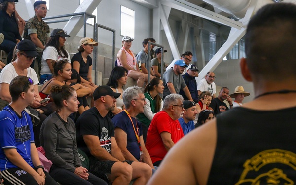 A crowd of civilians and military personnel in camouflage uniforms are seated on concrete bleachers or steps, watching an event off-camera. A muscular individual in a black sleeveless shirt with a yellow military unit insignia stands in the foreground with his back to the camera. The setting appears to be an indoor or covered military facility or arena.