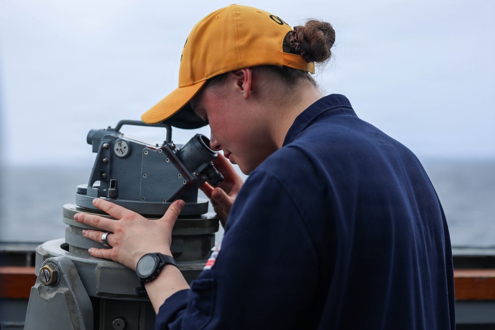A young woman in a navy blue uniform and yellow baseball cap is using a gyrocompass or pelorus (bearing circle) aboard a naval or coast guard vessel. She is looking through the sighting vane of the instrument, likely taking a navigational bearing. The overcast sky and open water visible in the background confirm an at-sea setting.