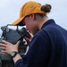 A young woman in a navy blue uniform and yellow baseball cap is using a gyrocompass or pelorus (bearing circle) aboard a naval or coast guard vessel. She is looking through the sighting vane of the instrument, likely taking a navigational bearing. The overcast sky and open water visible in the background confirm an at-sea setting.