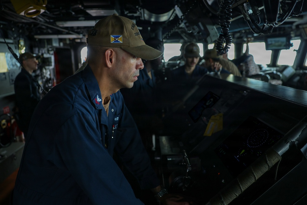The image shows U.S. Navy personnel on the bridge of a naval vessel, likely a destroyer based on the 'DDG' designation visible on the cap of the foreground sailor. The primary subject is a male sailor in dark blue coveralls and an olive/tan ball cap, focused on navigation or control equipment at a console. Several other crew members are visible in the background, also stationed at various bridge positions.