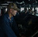 The image shows U.S. Navy personnel on the bridge of a naval vessel, likely a destroyer based on the 'DDG' designation visible on the cap of the foreground sailor. The primary subject is a male sailor in dark blue coveralls and an olive/tan ball cap, focused on navigation or control equipment at a console. Several other crew members are visible in the background, also stationed at various bridge positions.
