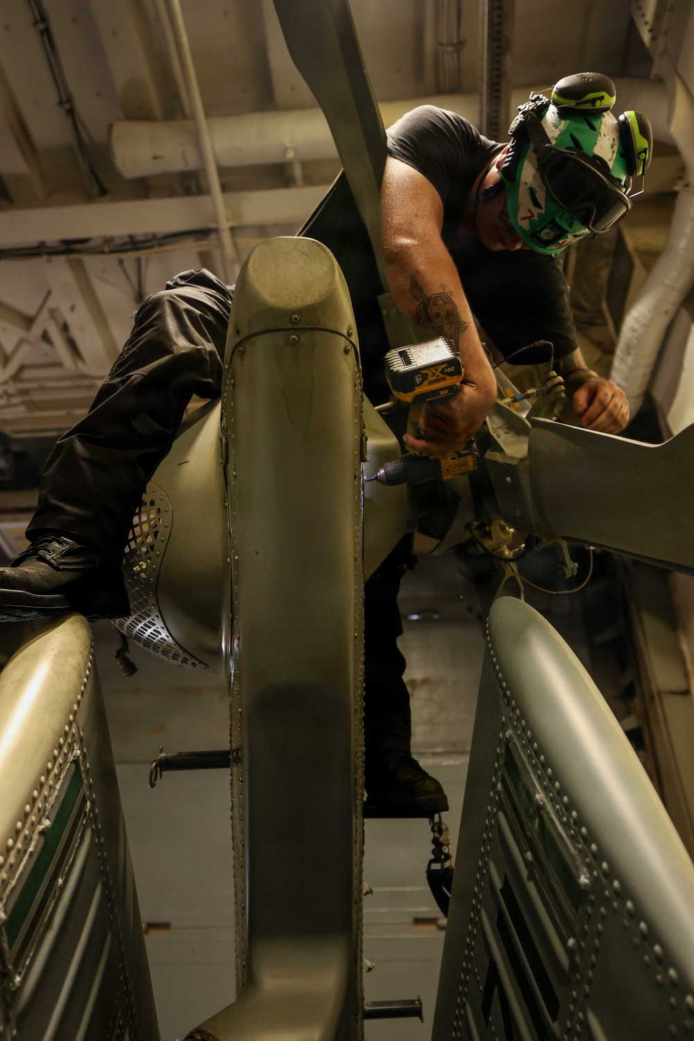 A technician or mechanic is performing maintenance work on what appears to be a military helicopter, likely in a hangar bay. The worker is positioned atop or between rotor blade assemblies, using a cordless power drill. The setting appears to be an enclosed hangar, possibly aboard a naval vessel given the overhead pipe and structural configurations.