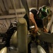 A technician or mechanic is performing maintenance work on what appears to be a military helicopter, likely in a hangar bay. The worker is positioned atop or between rotor blade assemblies, using a cordless power drill. The setting appears to be an enclosed hangar, possibly aboard a naval vessel given the overhead pipe and structural configurations.