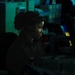 A person wearing a headset is seated at a workstation in a dimly lit, multi-monitor operations environment. The scene is illuminated primarily by blue-green ambient lighting, consistent with a command center, naval combat information center, or similar operational facility. At least one additional person is visible in the background at a separate workstation.