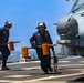 Two naval deck crew members in blue coveralls and safety gear are carrying orange wheel chocks on a ship's flight deck. They are working near a military helicopter, visible on the right side of the frame, in what appears to be a deck operations or aircraft handling procedure. The scene takes place at sea under clear blue skies.