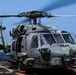 A U.S. Navy helicopter, numbered '707', is positioned on the flight deck of a naval vessel at sea with its rotors spinning. The aircraft appears to be an MH-60R Seahawk, identifiable by its distinctive nose-mounted sensors and gray naval paint scheme. The helicopter is chained to the deck and appears to be either preparing for takeoff or having just landed.