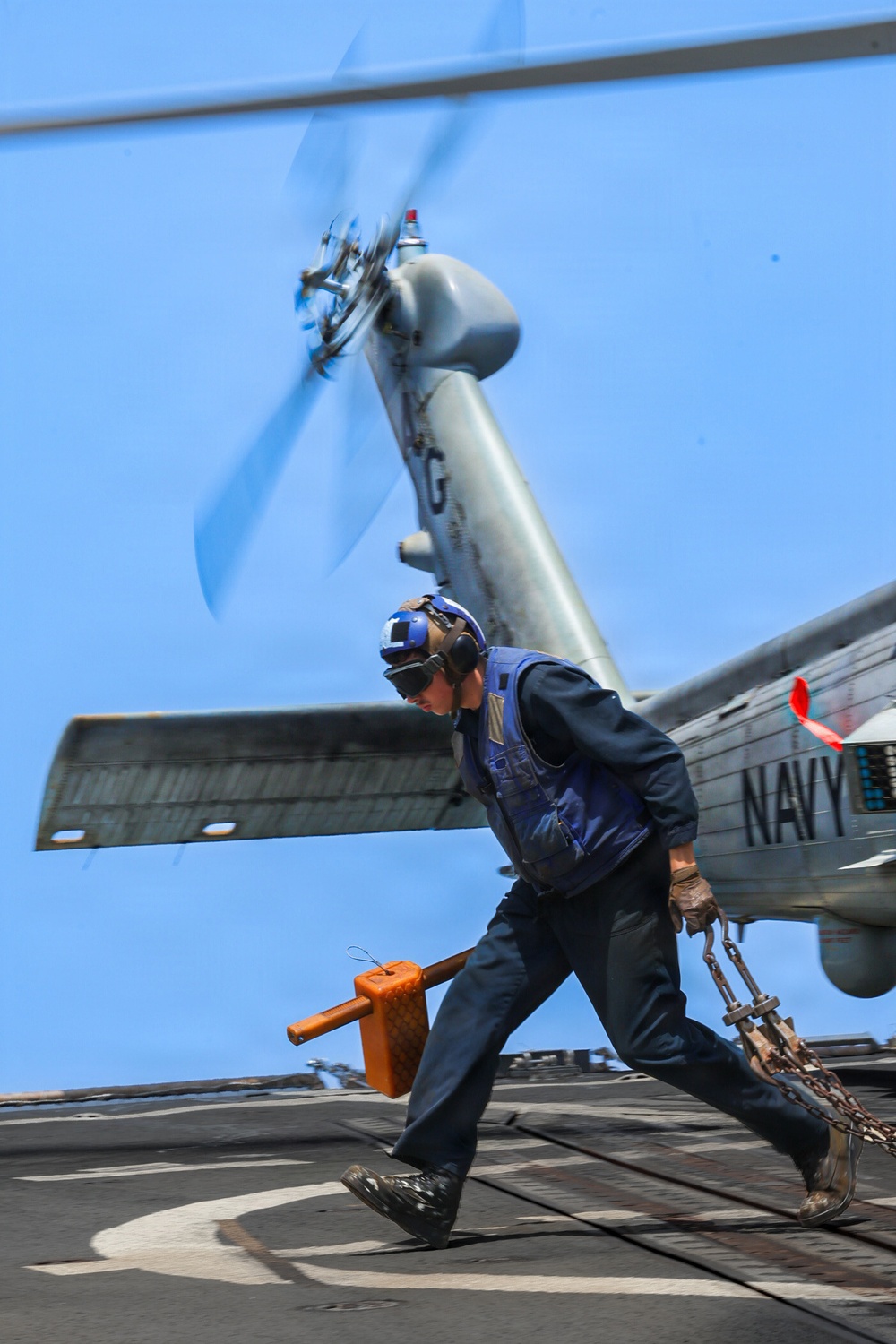 A U.S. Navy deck crew member is running across a ship's flight deck while a helicopter with spinning rotors is present overhead. The sailor is carrying tie-down chains and an orange chock, indicating active aircraft handling operations. The scene captures the dynamic and hazardous environment of carrier or naval vessel flight deck operations.