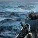 A naval vessel is conducting an underway replenishment (UNREP) operation in rough sea conditions. A group of sailors wearing colored hard hats and blue coveralls are stationed along the ship's deck, monitoring a fuel or supply line extended over the churning ocean. The operation appears to be in progress with rigging, hoses, and tensioned lines visible between ships.