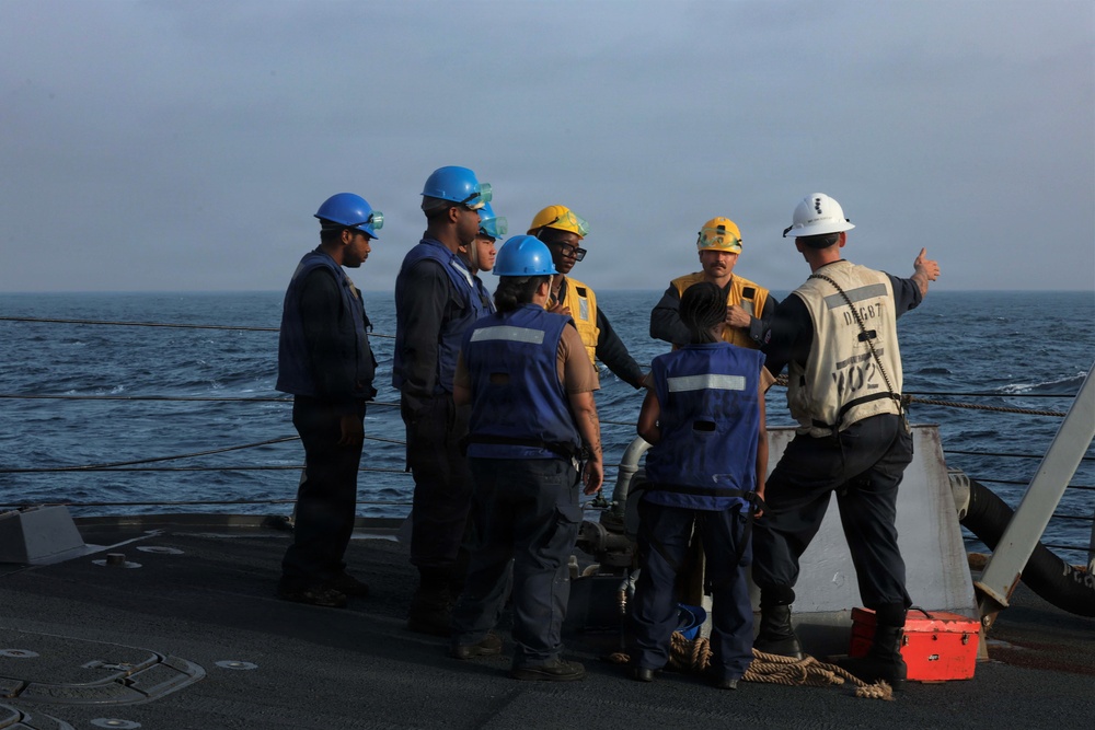 A group of naval personnel are gathered on the deck of a warship at sea, appearing to receive a briefing or instructions from a senior officer wearing a white hard hat. The personnel are wearing various colored hard hats (blue, yellow, white) and safety vests, indicating different roles or departments. The ship identifier 'DDG-87' is partially visible on the back of one crew member's vest.