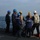 Routine Operations Aboard USS Mason (DDG 87) A group of naval personnel are gathered on the deck of a warship at sea, appearing to receive a briefing or instructions from a senior officer wearing a white hard hat. The personnel are wearing various colored hard hats (blue, yellow, white) and safety vests, indicating different roles or departments. The ship identifier 'DDG-87' is partially visible on the back of one crew member's vest.