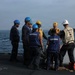 A group of naval personnel are gathered on the deck of a warship at sea, appearing to receive a briefing or instructions from a senior officer wearing a white hard hat. The personnel are wearing various colored hard hats (blue, yellow, white) and safety vests, indicating different roles or departments. The ship identifier 'DDG-87' is partially visible on the back of one crew member's vest.