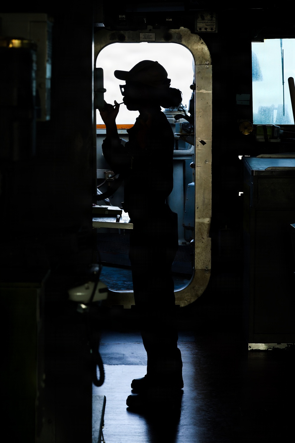 A silhouetted figure stands in a doorway or hatchway aboard what appears to be a naval or military vessel, backlit by an overcast sky outside. The person is wearing a cap and appears to be holding a communication device or radio to their face. The image is a dramatic high-contrast silhouette photograph taken from inside the ship's interior.