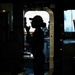 A silhouetted figure stands in a doorway or hatchway aboard what appears to be a naval or military vessel, backlit by an overcast sky outside. The person is wearing a cap and appears to be holding a communication device or radio to their face. The image is a dramatic high-contrast silhouette photograph taken from inside the ship's interior.