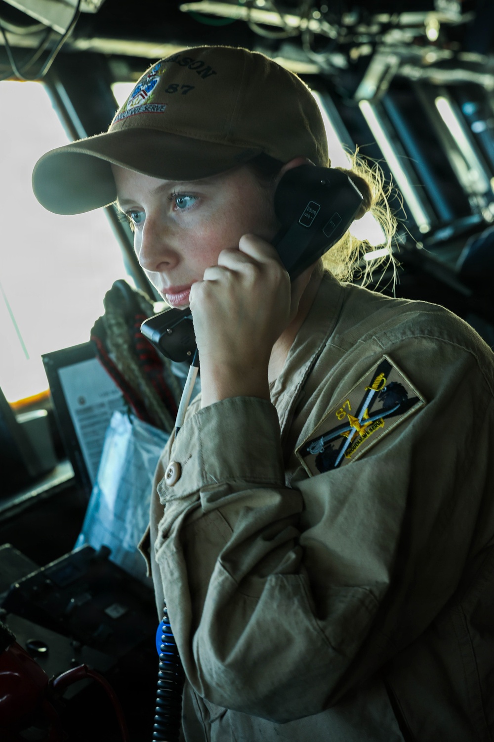 A female military service member is stationed on what appears to be a naval vessel bridge, speaking into a sound-powered telephone handset. She is wearing a tan/khaki uniform coverall and a matching ball cap with a unit patch visible, bearing the number '87'. The setting is the interior of a ship's bridge or combat information center, with navigation equipment visible in the background.