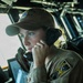 A female military service member is stationed on what appears to be a naval vessel bridge, speaking into a sound-powered telephone handset. She is wearing a tan/khaki uniform coverall and a matching ball cap with a unit patch visible, bearing the number '87'. The setting is the interior of a ship's bridge or combat information center, with navigation equipment visible in the background.